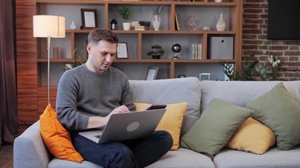 男人坐在笔记本电脑上，坐在沙发上在家工作。男性使用计算器笔记本电脑应用程序计算成本，通过电子银行支付应用。业务、家庭开支、预算管理. — 图库视频影像