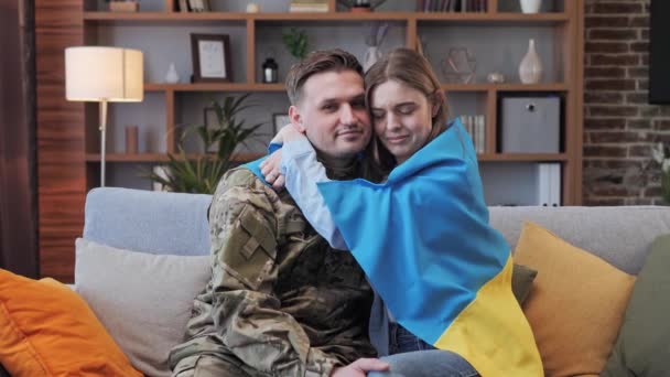 Soldado en uniforme militar y su chica envuelta en la bandera de Ucrania se abrazan en casa en el sofá. Las hostilidades militares. Movilización de la población. Russias atacan Ucrania. Detener la guerra. — Vídeos de Stock