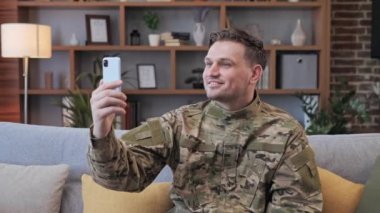 Mutlu pozitif asker evinde koltukta oturuyor ve ailesiyle iletişim kurmak için internet uygulamalı cep telefonunu kullanıyor. Adam savaştan sonra eve döndü. Özgür bağımsız insanlar.