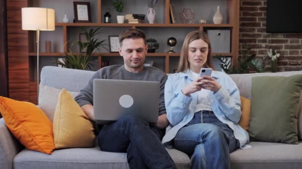 若い幸せなカップルは自宅でラップトップとスマートフォンを使用して楽しんでいます。女の子と男が話して、笑って、ラップトップショッピングの画面を見て、オンラインで配達を注文し、ソーシャルメディアアプリをチェックする. — ストック動画