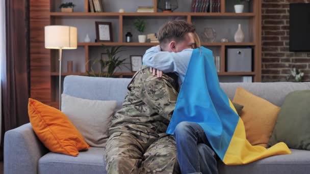 Manžel ve vojenské uniformě a jeho žena zabaleni v vlajce Ukrajiny objímají doma na gauči. Vojenské nepřátelství. Mobilizace populace. Rusové útočí na Ukrajinu. Zastavte válku.. — Stock video