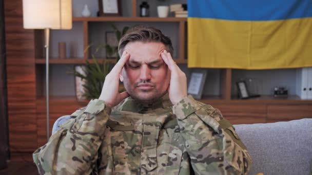 L'uniforme militare del soldato si siede sul divano e ha un forte mal di testa. Bandiera ucraina sullo sfondo. La Russia invade un paese democratico europeo. Fermate la guerra. Depressione. — Video Stock