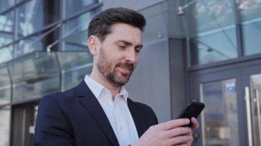 Klasik Takım Elbiseli Başarılı İş Adamının Portresi Smartphone Outdoor 'unu kullanıyor. Yakışıklı erkek girişimci cam ofis binasının yanında dikiliyor. Çekici bir şekilde gülümsüyor. İş için. - İnsanlar. Finans.