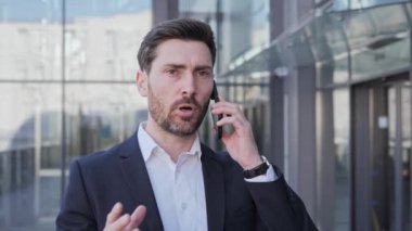 Klasik takım elbiseli endişeli iş adamı dışarıda cep telefonuyla konuşuyor. İş sorularıyla ilgili kötü haberleri olan yakışıklı sinirli bir adam. Elleriyle işaret ediyor. Ofisin yakınındaki hoşnutsuzluğunu gösteriyor..