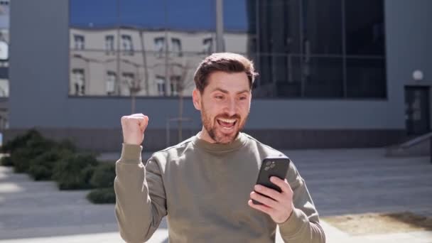 都市部のスマートフォンの画面を見て興奮したビジネスマン。幸せな人は屋外の携帯電話で良いニュースを読む。近代的なガラスの建物の近くで勝利を祝う驚きのビジネスマン. — ストック動画