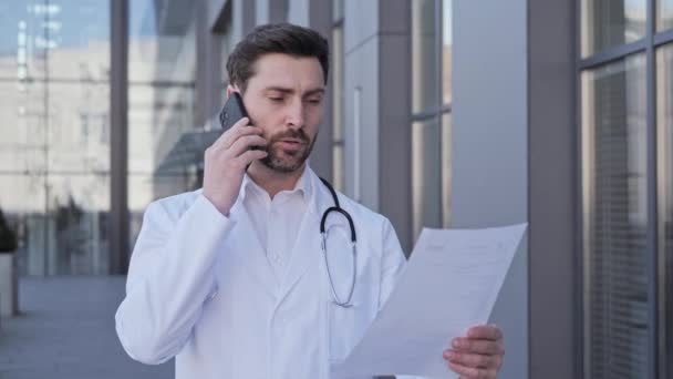 Врач со стетоскопом держит отчет о состоянии здоровья пациентов и общается с ним по мобильному телефону. Врач-терапевт держит лист бумаги с выводом заболевания. — стоковое видео