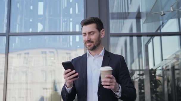 Neşeli girişimci yakışıklı sakallı adam şehirde cep telefonuyla konuşuyor kahve içiyor dışarıda cep telefonuyla iş haberleri okuyor. Teknoloji ve iletişim konsepti. — Stok video