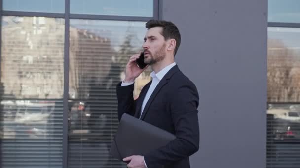 세련 된 양복을 입은 잘생긴 사업가가 직장 문제를 해결하는 동료와 휴대폰으로 온라인 대화를 나누는 동안 문서가 담긴 폴더를 들고 있다. 카메라의 측면 사진. — 비디오