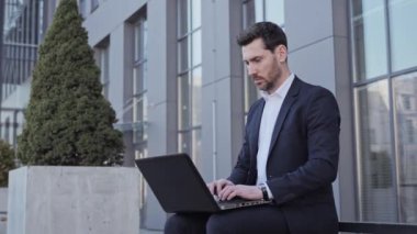 Modern ofis binasının yakınındaki bankta çalışırken dizüstü bilgisayar klavyesinde daktilo eden konsantre olmuş, sakallı iş adamının yakın plan fotoğrafını çek.. 