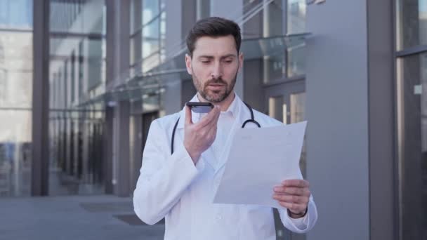 Steteskoplu bir erkek doktor, elinde hastaların tıbbi geçmişi ile ilgili bir rapor tutuyor. Ses tanıma mesajını hoparlörden kaydediyor.. — Stok video