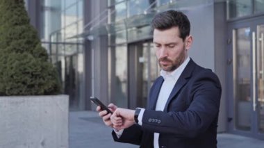 Başarılı bir iş adamı resmi olarak akıllı telefon hızlı daktilo kullanarak iş yerine e-posta göndermeye vakit bulamıyor. Kendine güvenen genç girişimci ofis binasının yakınındaki saatine bakıyor.