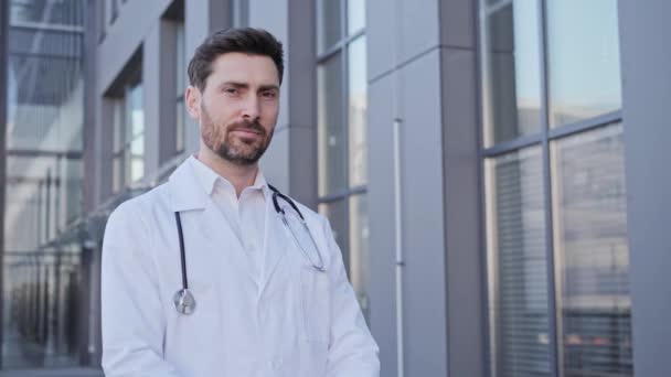 Retrato de un joven atractivo que da una sonrisa dentada en uniforme de médicos con un estetoscopio Un profesional médico masculino seguro sonriendo a la cámara mientras dobla las manos fuera del hospital. — Vídeos de Stock