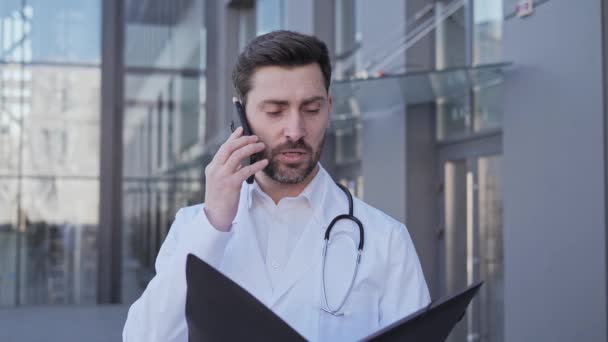 Médecin thérapeute professionnel détient un dossier avec un rapport sur la maladie des patients parlant sur un téléphone portable avec un patient malade. Consultation médicale masculine en ligne par téléphone près d'une clinique moderne. — Video