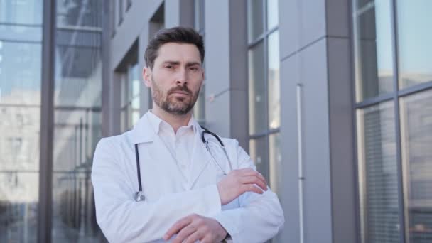 Portrait d'un médecin caucasien homme travailleur de la santé debout à l'extérieur de l'hôpital confiant regardant devant la caméra. Visage du chirurgien portant des gommages et un stéthoscope posant sur fond de clinique. — Video