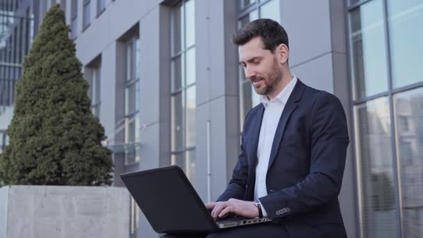 Lächelnd tippt ein gutaussehender, bärtiger Geschäftsmann in formeller Kleidung auf der Tastatur eines Laptops, während er auf einer Bank im Freien in der Nähe eines modernen Bürogebäudes online arbeitet. Erfolgreiche Menschen. — Stockvideo