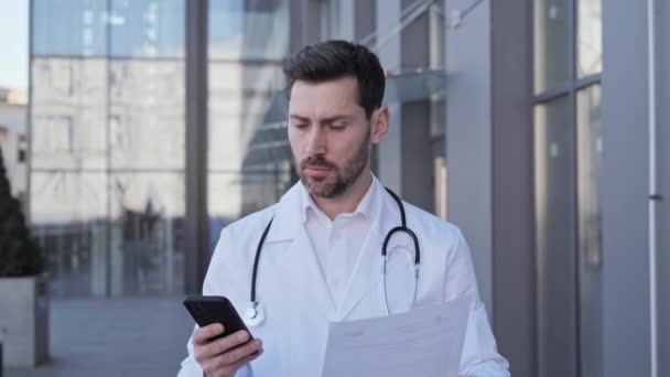 Medico professionista maschile medico con uno stetoscopio digitando chat invio di messaggi online. Terapista tiene un foglio di carta con la conclusione della malattia sullo sfondo della clinica moderna. — Video Stock