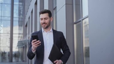 Akıllı telefon uygulaması kullanarak şık takım elbiseli, gülümseyen sakallı adam. Yakışıklı işadamı tomarla tomarla internet arıyor. Ofis bölgesinde yürüyor. Belgelerle birlikte şehir caddesinde klasör tutuyor..