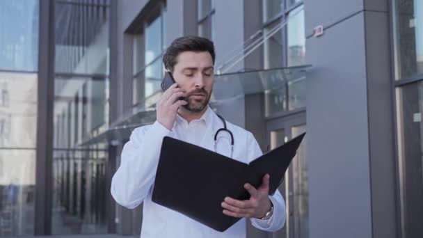 Professional doctor therapist the doctor holds a folder with medical documents and consults the patient by phone explains the course of the disease for further treatment near a modern clinic. — Stock Video