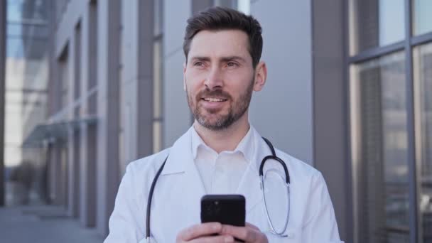 Médecin adulte debout près de la clinique à l'extérieur tenant un message texte de téléphone portable, écran défilant du smartphone. Médecin d'âge moyen envoie une prescription en ligne au patient pendant une pandémie. — Video