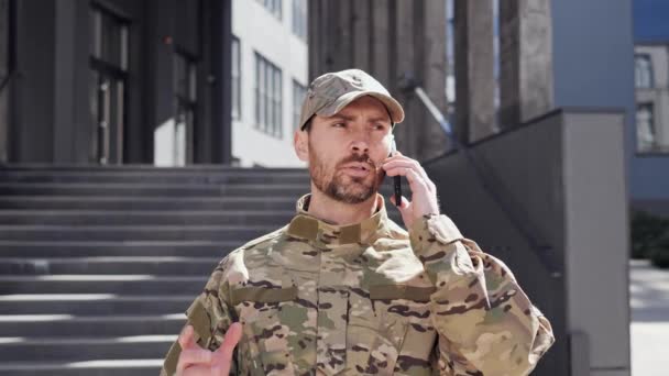 Army soldier in a cap having online mobile phone call with his superiors outdoors. The soldier gestures with hands during the conversation looks confident and focused standing near modern building. — Stock Video