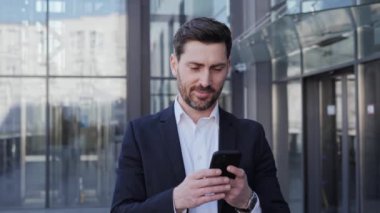 Başarılı Tatmin Olmuş Takım Elbiseli İşadamı 'nın portresi açık havada akıllı telefonunu kullanıyor. Yakışıklı erkek girişimci cam ofis binasının yanında durmuş sohbet ediyor ve mesaj gönderiyor. İş dünyası. 