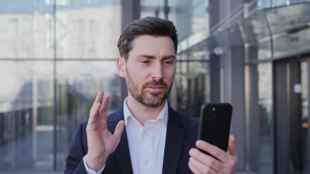 Hombre de negocios serio haciendo video chat en línea llamada de conferencia utilizando la aplicación de teléfono móvil se ve enfocado y confiado fuera de la oficina de pie en la calle urbana de la ciudad. Gente de negocios tecnología digital. — Vídeo de stock