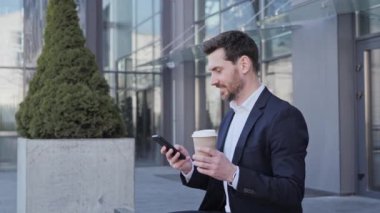 Kendine güveni tam bir iş adamı büyük modern iş bölgesinin yakınında oturuyor. Genç sakallı adam Smartphone 'da mesaj atıyor ve dinlenmek için kahve içiyor. Başarı. - İnsanlar. Vay be. Uygulamalar.