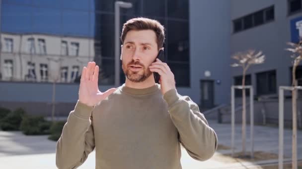 Hombre barbudo joven de buen aspecto con aspecto elegante caminando por la calle del centro de la ciudad hablando en el teléfono móvil gestos emocionales con las manos. Cool chico con estilo camina conversaciones en el teléfono inteligente. — Vídeo de stock