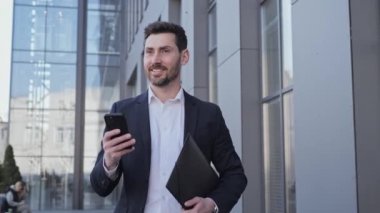 Akıllı telefon uygulaması kullanarak şık takım elbiseli yakışıklı bir adam. Yakışıklı işadamı tomarla tomarla internet arıyor. Ofis bölgesinde yürüyor. Belgelerle birlikte şehir caddesinde klasör tutuyor..