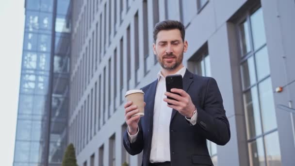 Joven Hombre Profesional Chateando Enviando sms trabajando en su Smartphone. Escribir un mensaje. De pie cerca del Gran Edificio Industrial. Beber café para llevar. Chaqueta clásica. Aplicaciones. Gente de negocios. — Vídeo de stock