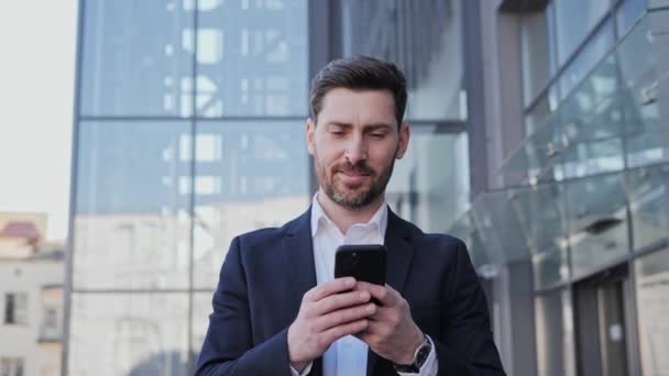 Счастливый кавказский молодой предприниматель просматривает мобильное приложение данных телефона в интернете во время офисного перерыва, гуляя по деловому району. Успешный бизнесмен идет на работу. Деловые люди. Успех. — стоковое видео