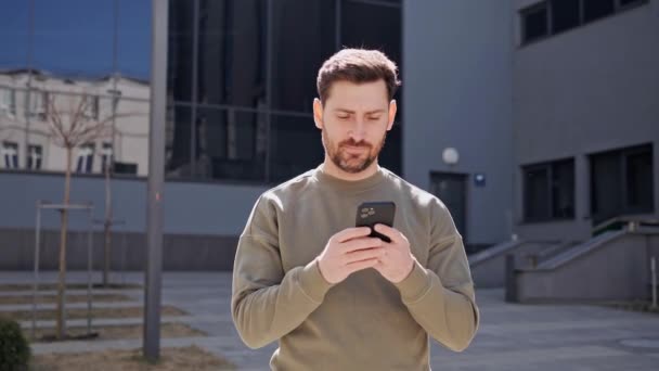 Handsome young businessman using a mobile phone and smiling. Sending text messages while standing near a modern office building. Wearing a casual clothes. Social network. Applications. Smartphone. — Stock Video