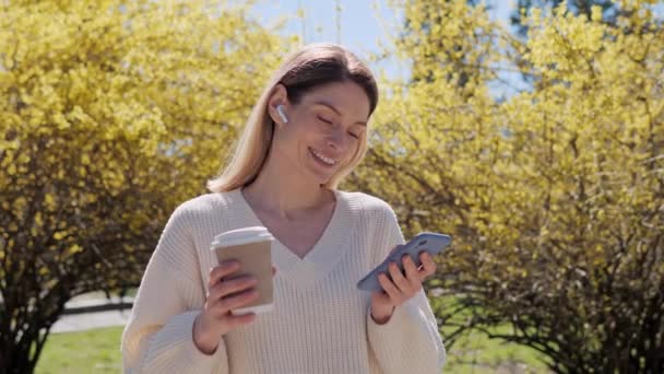 Χαμογελώντας όμορφη γυναίκα χρησιμοποιώντας το κινητό τηλέφωνο χτυπώντας οθόνη κύλισης του smartphone περιήγηση στο διαδίκτυο δεδομένα εφαρμογών κοινωνικής δικτύωσης κρατώντας ζεστό καφέ στο πάρκο κοντά σε ένα δέντρο με κίτρινα φύλλα σε ζεστή ηλιόλουστη μέρα. — Αρχείο Βίντεο