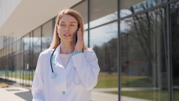专业女医生身穿白衣，带听诊器，在现代诊所附近有手机通话，在线咨询远程病人，远程解释治疗室外分享建议. — 图库视频影像