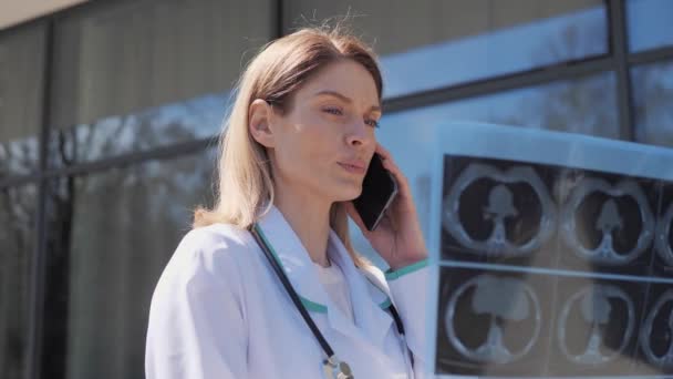 专业的、有经验的女医生学习X光图像的结果可以远程向病人提供建议。身穿白色外套和听诊器的妇女讲述了病人在室外接受X光检查的情况. — 图库视频影像