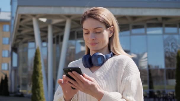 Bella donna sms messaggistica utilizzando una nuova applicazione per la navigazione smartphone internet digitando sms vestirsi in abiti casual con auricolari in piedi in un parco con un edifici sfocati in background. — Video Stock