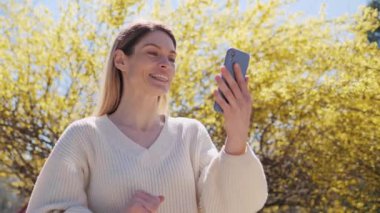Beyaz kazaklı, cep telefonu kullanan güzel bir kadın güneşli bir günde parkta sarı yapraklı bir ağacın yanında görüntülü konuşma yapıyor. Kadın konferans sohbeti yapıyor. Boş zaman geçiriyor..