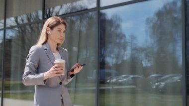 Şehir caddesinde yürürken cep telefonuyla sabah kahvesi içen orta yaşlı güzel bir iş kadını. Bir kadın ileriye bakıyor ve düşünceli bir şekilde uzaklaşıyor. Sosyal ağ. Akıllı telefonlar. Uygulamalar.