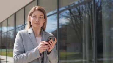 Kendine güvenen Dalgın İş Kadını büyük modern iş binasının yakınında sokakta yürüyor. Yetişkin Kadın Smartphone 'daki mesajları kontrol ediyor. Başarı. İş adamları. - Kadın. Uygulamalar.