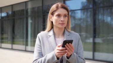 Şehir caddesinde duran mutlu iş kadını cep telefonuna bakıyor internette sohbet ediyor sosyal medyada düşünceli cevaplı mesajlar veriyor açık hava cihaz kullanıyor..
