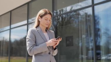 Gülümseyen yetişkin iş kadını iş bölgesinde duruyor. Telefon mesajları, kaydırmalı ekranlar, internet sosyal medyası, modern internet uygulamaları, internet uygulamaları, iletişim, güneşli gün başarısı kullanıyor..