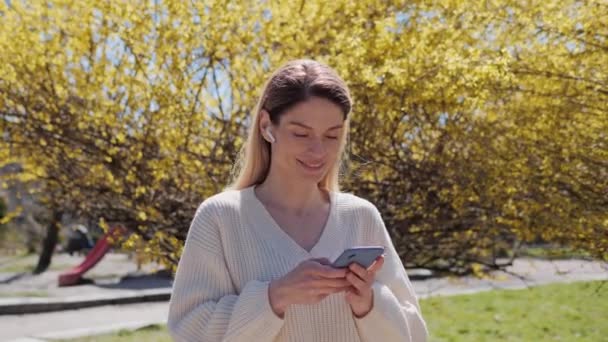 Senhora feminina em um suéter branco usando telefone celular menina tocando tela de rolagem de dados de aplicativos de mídia social de navegação de smartphones no parque perto de uma árvore com folhas amarelas em um dia ensolarado quente. — Vídeo de Stock