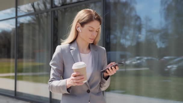 Beautiful young businesswoman in a formal suit holding coffee and using a modern smartphone app standing outdoors, a professional female employer typing a text message on a mobile phone outside. — 비디오