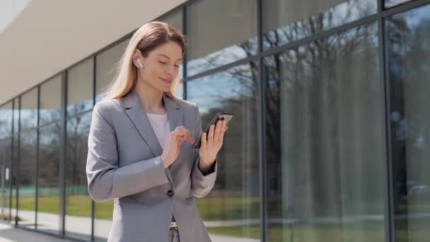 Handsome young businesswoman using a mobile phone and wireless headphones. Sending text messages while standing near a modern office building. Social network Applications Smartphone Business People. — 비디오