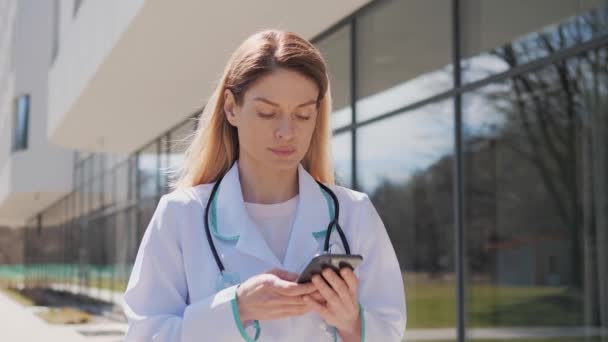 Female doctor physician with a stethoscope standing near clinic outdoors holding mobile phone texting message scrolling screen of smartphone sends online prescription to the patient during a pandemic. — стоковое видео