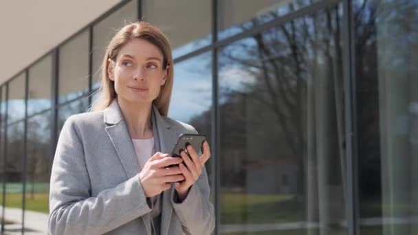 Confident Pensive Businesswoman walking on the street near big modern business building. Adult Woman texting Messages on Smartphone scrolling checking mail. Success. Business people. Woman. Apps. — Stock Video