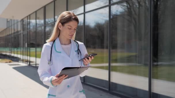 Ernstige vrouwelijke arts met smartphone typen bericht of antwoord aan de patiënt op de achtergrond van de moderne kliniek. Vrouwelijke arts in witte jas afluisteren op mobiele telefoon. Beroepsbeoefenaar. — Stockvideo