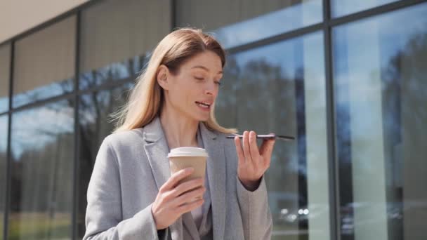 Bizneswoman wysyłając wiadomość głosową na smartfonie trzymając poranną kawę w pobliżu nowoczesnego budynku biurowego rozmawiając z asystentem mobilnym. Bizneswoman w formalnym garniturze za pomocą aplikacji na telefon komórkowy. — Wideo stockowe