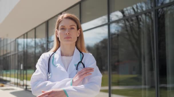 Retrato de doctora terapeuta de mediana edad vestida con bata médica con estetoscopio. Médico de confianza cruzando brazos mirando la cámara de pie cerca de la clínica moderna. Médico femenino enfocado posando. — Vídeo de stock