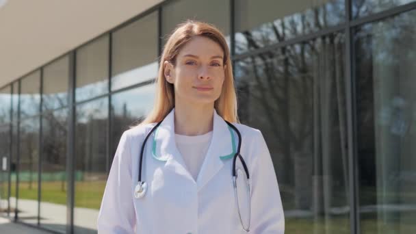 Close up retrato de branco médico trabalhador de saúde do sexo feminino em pé fora do hospital virando a cabeça sorrindo. Rosto confiante de cirurgião vestindo esfregaços e estetoscópio posando na câmera. Emoção real. — Vídeo de Stock